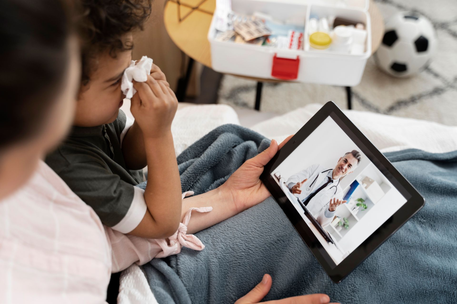 Person on couch using a tablet while another wipes tears; a stuffed soccer ball sits nearby.