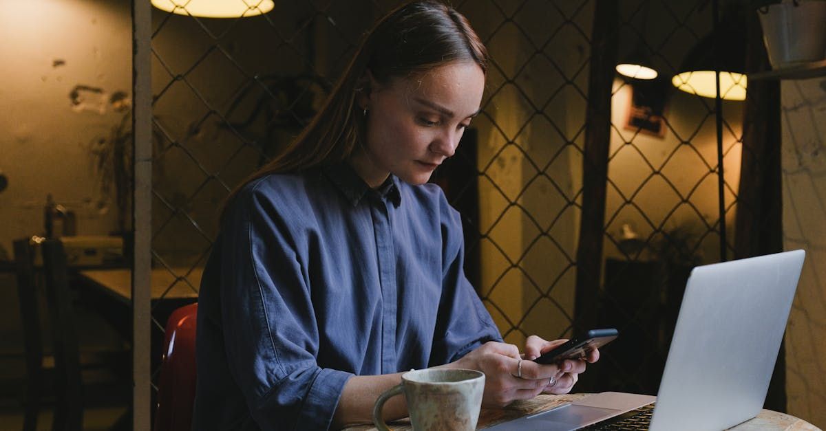 Woman in blue shirt using phone, sitting at a table with a laptop and coffee cup in a dim cafe.