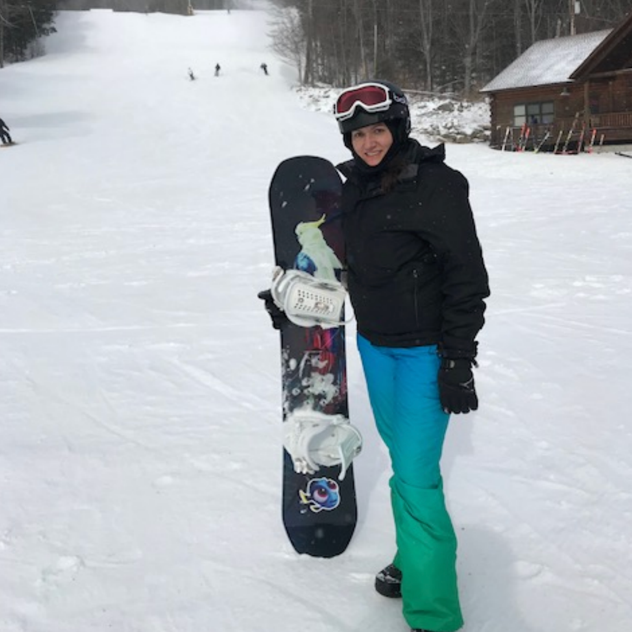 Person in black jacket and blue-green pants holding a snowboard on a snowy ski slope near a lodge.