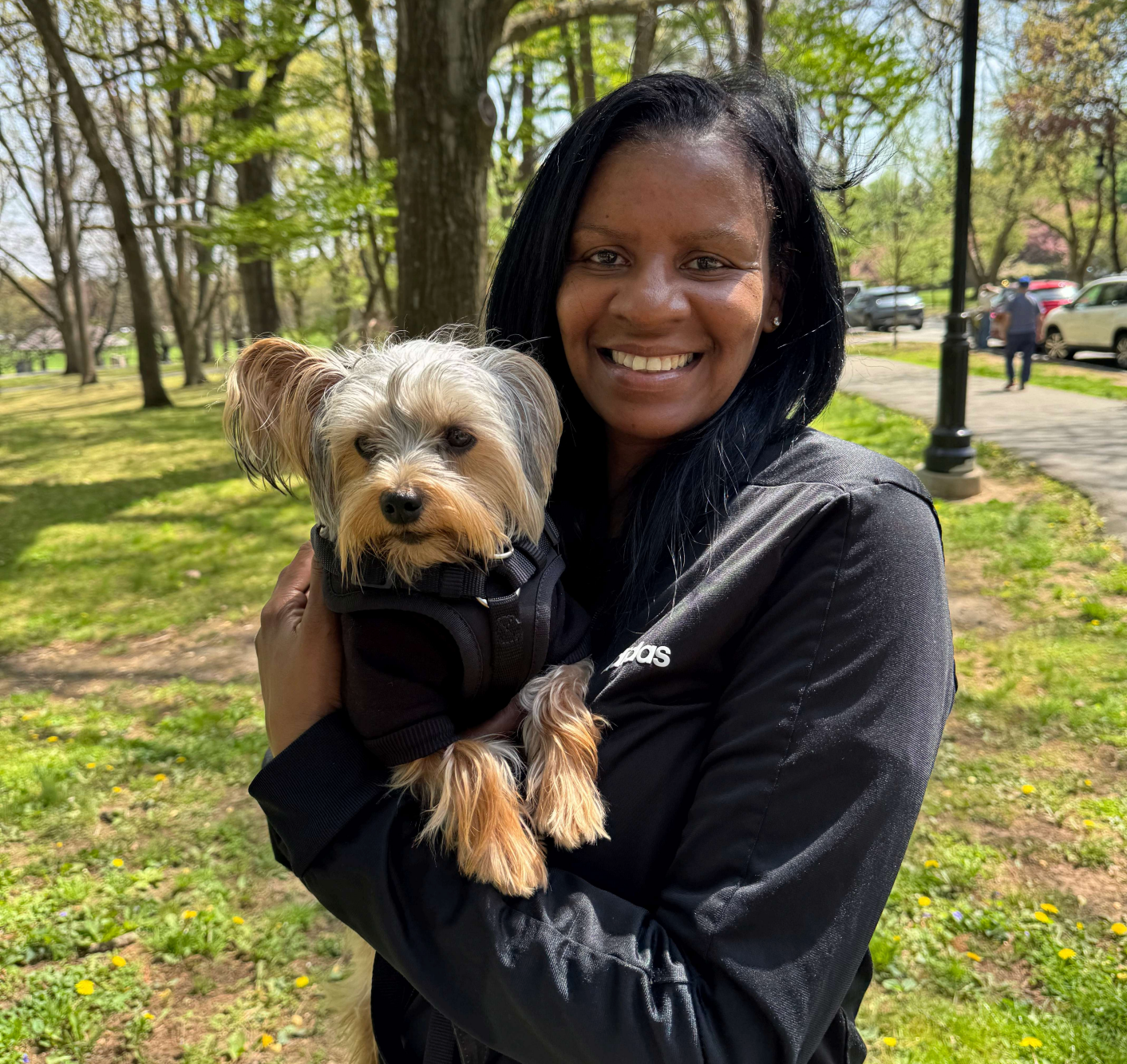 Woman smiling and holding a small dog in a sunny park with trees and grass