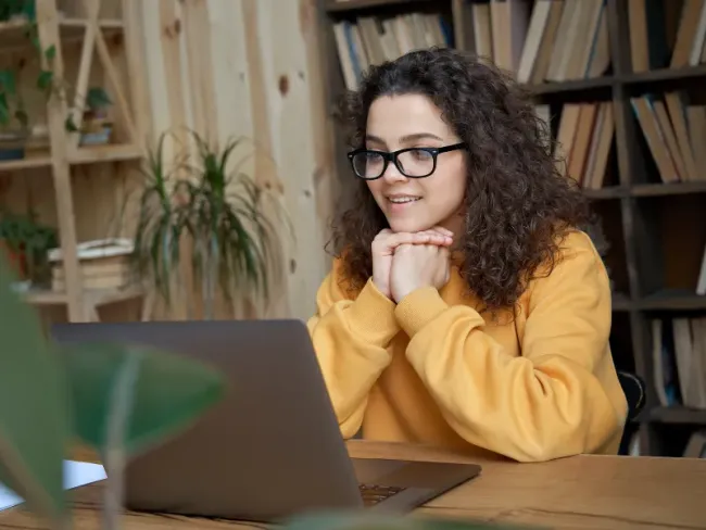 Woman with curly hair and glasses smiles at laptop, resting chin on hands, in a library.