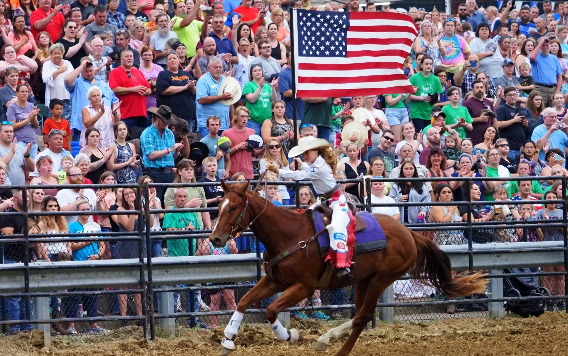 Cowgirl riding a brown horse in an arena, with a large American flag and cheering crowd behind her