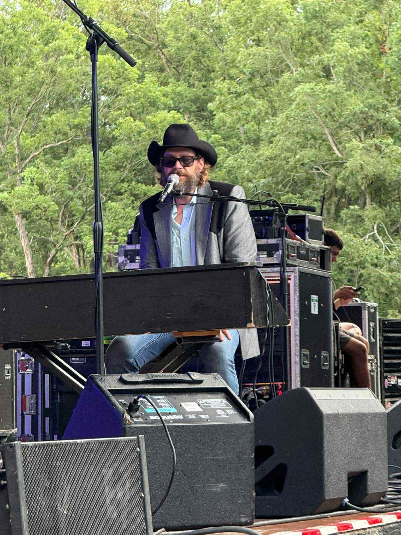 Musician at keyboard on outdoor stage, wearing black hat and sunglasses, with trees in the background
