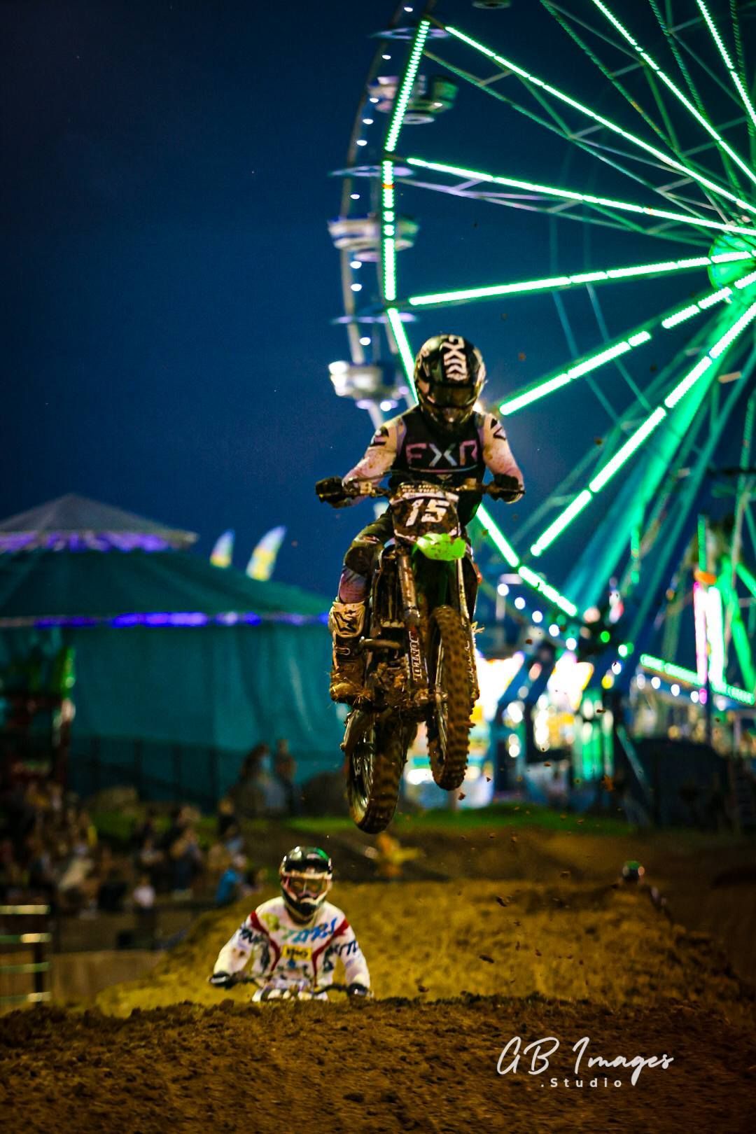 Motocross rider airborne at night in front of a glowing Ferris wheel at a fairground