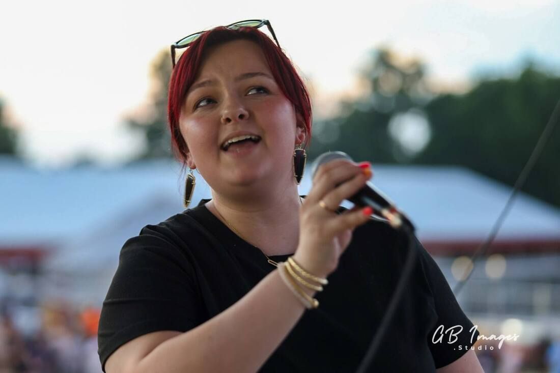 Singer holding a microphone outdoors, wearing black and smiling during a performance.