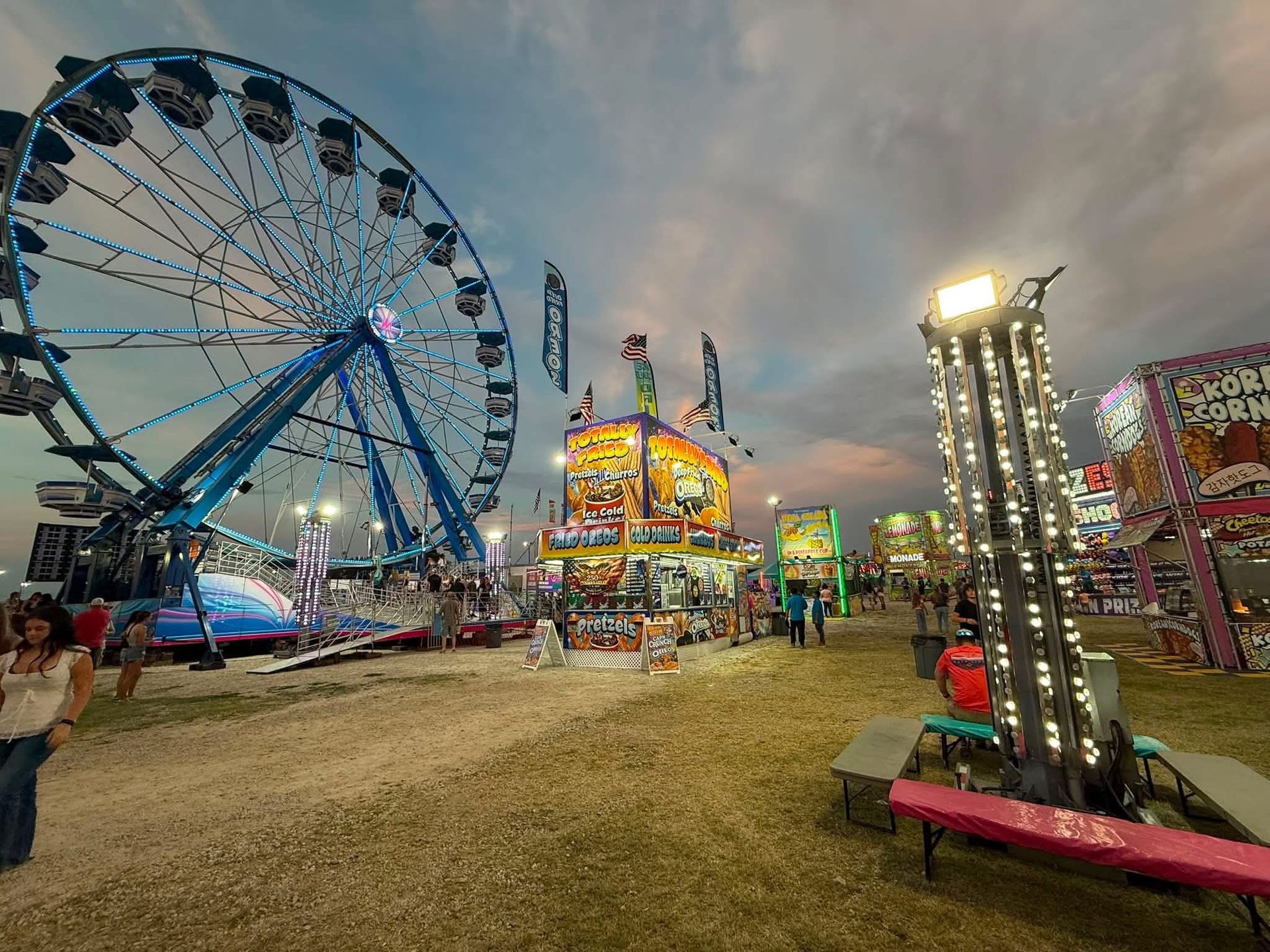 Crowded carnival midway with colorful food booths, bright banners, and people walking under a blue sky