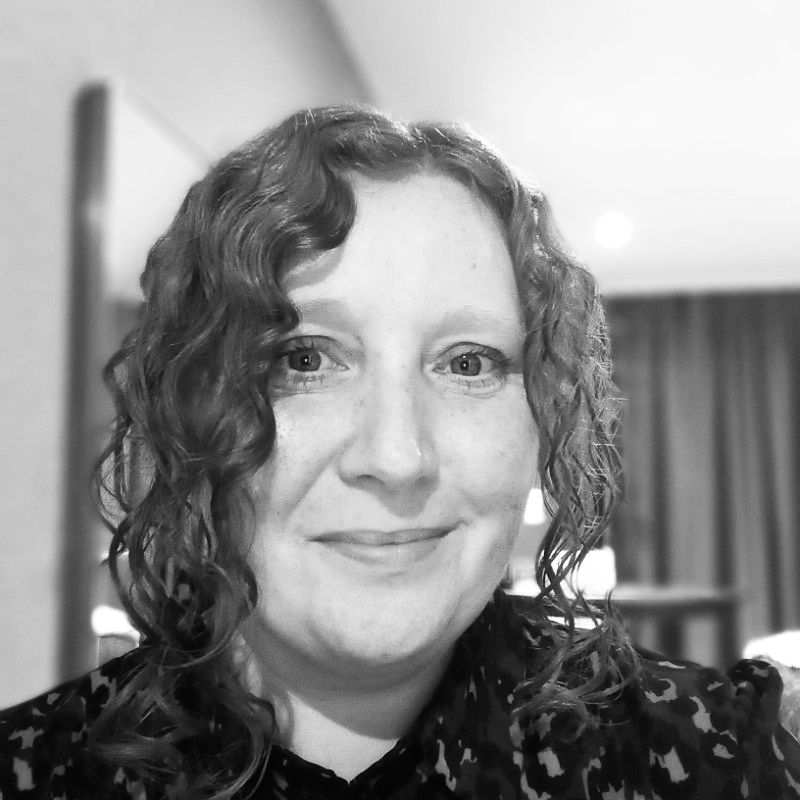A black-and-white selfie of a smiling person with shoulder-length curly hair wearing a patterned top in an indoor setting.