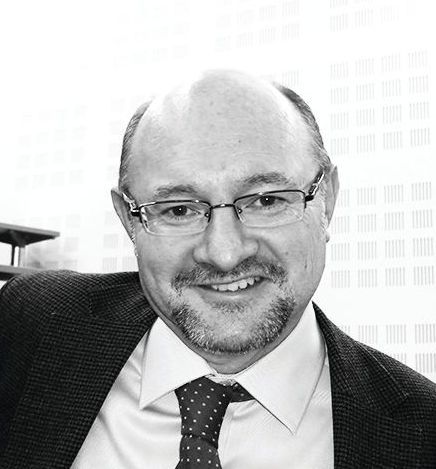 A smiling man wearing glasses, a white collared shirt, a dotted tie, and a dark blazer.