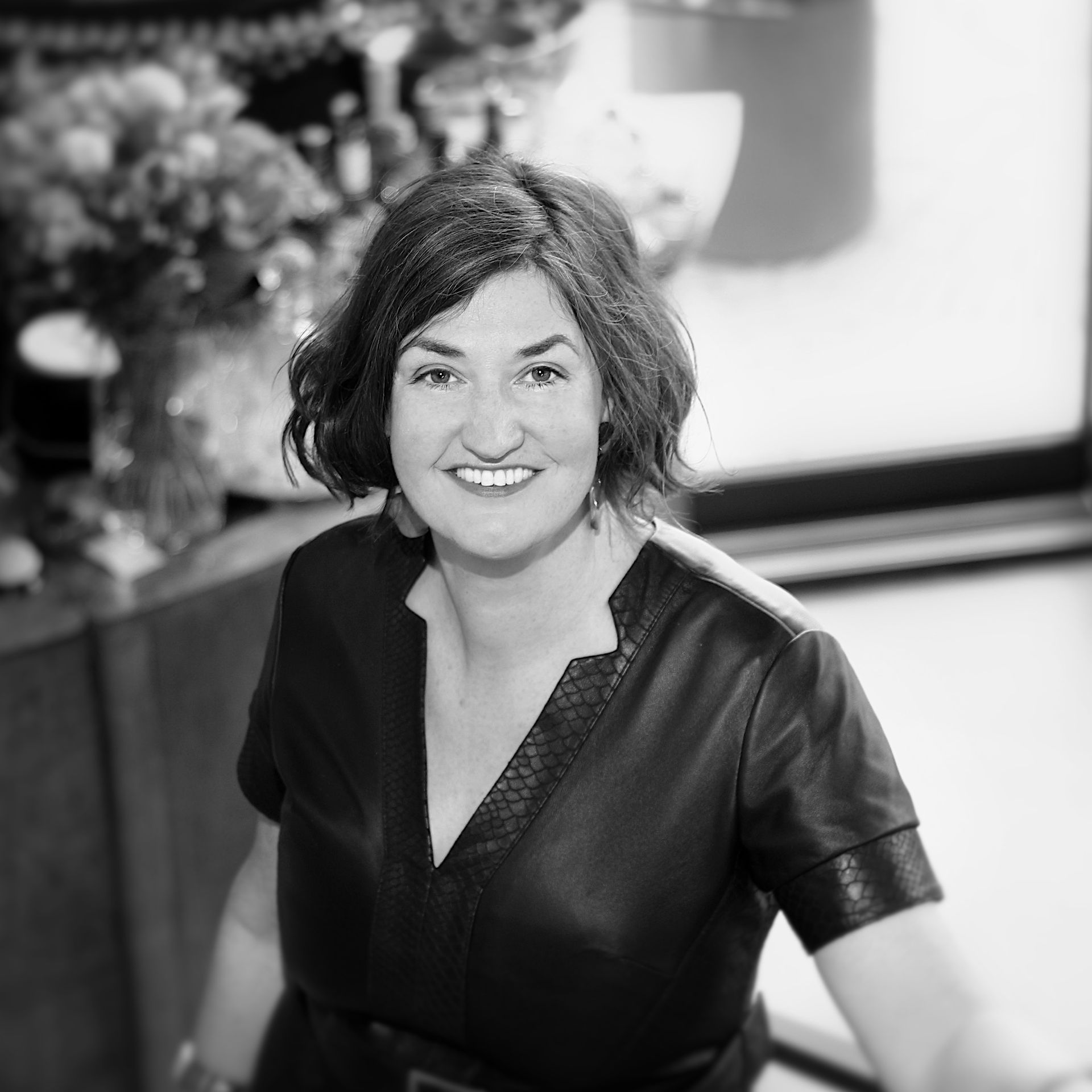 A professional portrait of a smiling person with shoulder-length wavy hair, wearing a dark v-neck top in an indoor setting.