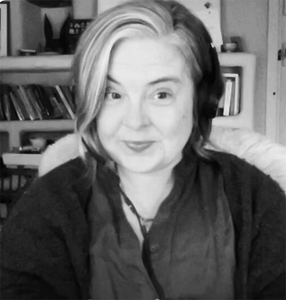A black-and-white portrait of a person with shoulder-length hair, wearing a dark shirt, sitting in front of bookshelves.