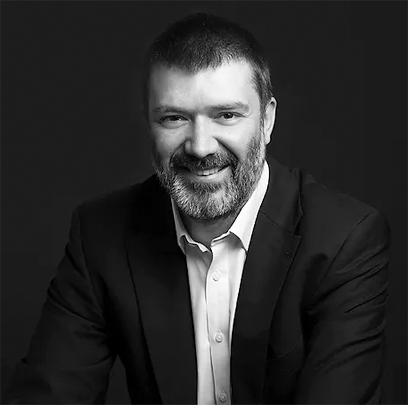 Black-and-white headshot of a smiling person with short hair and a beard, wearing a suit jacket and dress shirt.