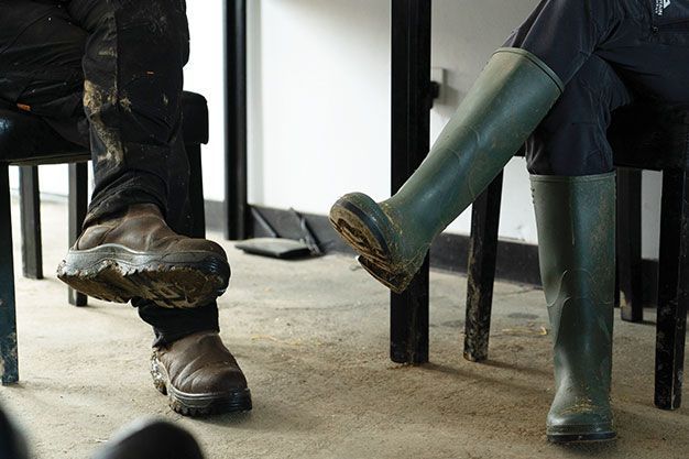 Two people sitting in chairs, one wearing brown work boots and muddy pants, the other in olive green knee-high rubber boots.