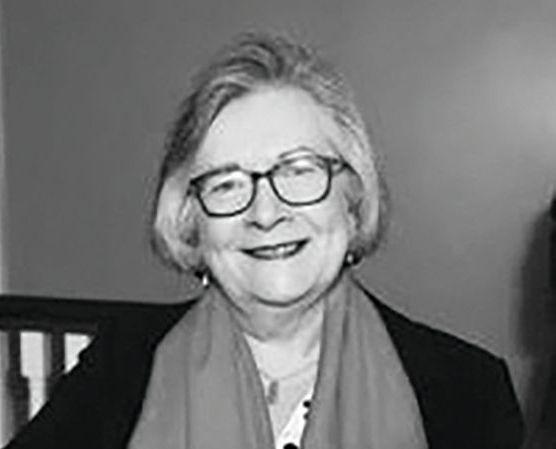 A person with short hair and glasses smiles warmly, wearing a jacket and scarf in a portrait shot.
