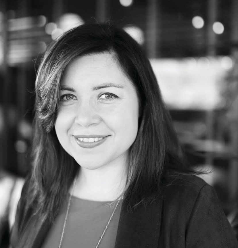 A black-and-white headshot of a smiling person with shoulder-length dark hair, wearing a blazer over a collared top.