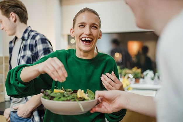 A smiling person in a green sweater holds a bowl of salad while interacting with others in an indoor setting.