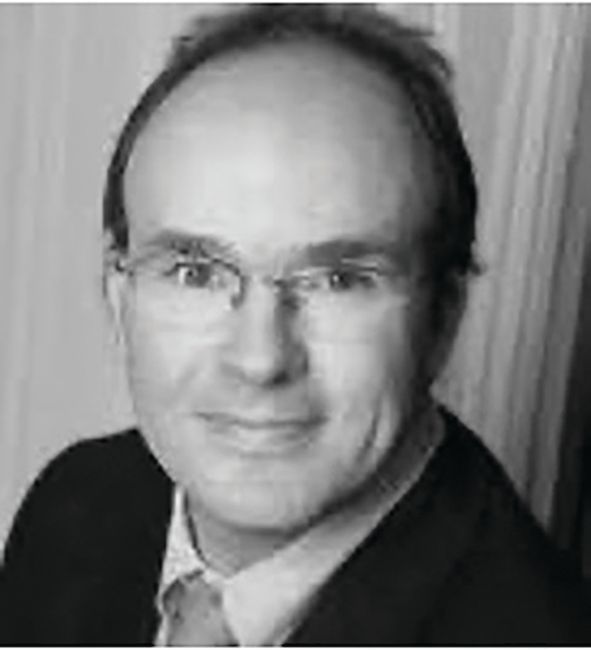 Head-and-shoulders portrait of a person wearing glasses and a suit, looking at the camera against a vertical-blind backdrop.