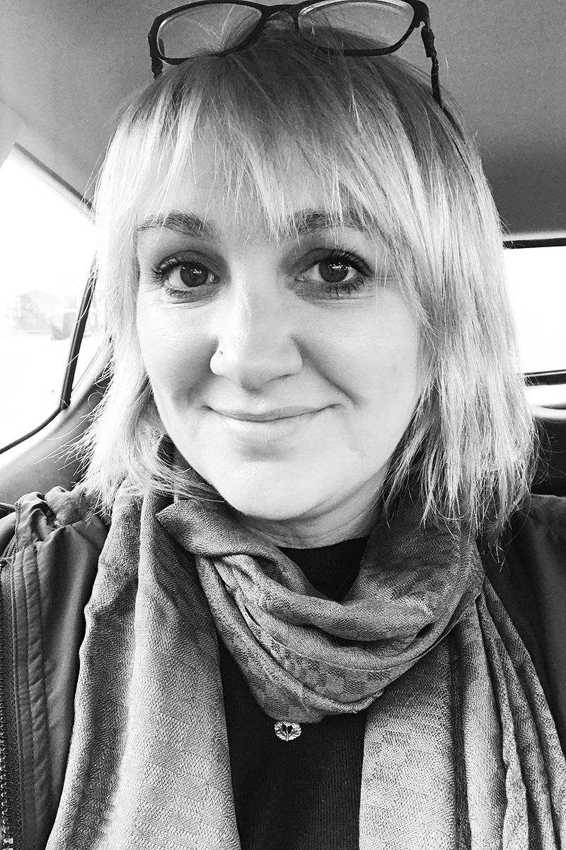 A black and white selfie of a person smiling, wearing a scarf and glasses perched on their head inside a vehicle.