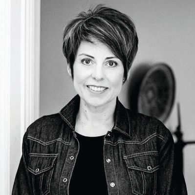 A black-and-white portrait of a person with a short, layered hairstyle, smiling while wearing a denim jacket.
