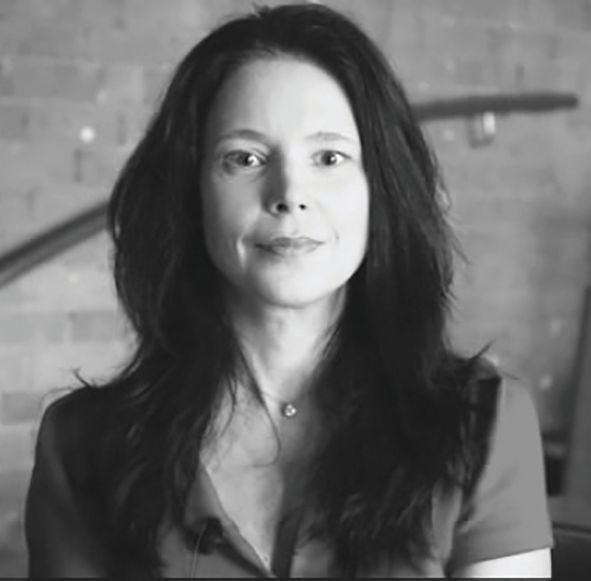 Black-and-white headshot of a woman with long dark hair, wearing a simple necklace, against a textured brick wall.