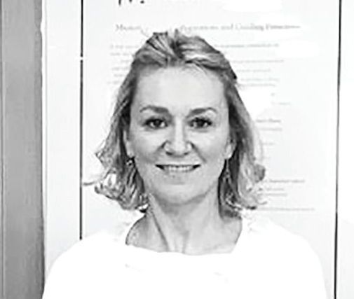 A person with shoulder-length hair smiling at the camera, positioned in front of a white wall with indistinct text.