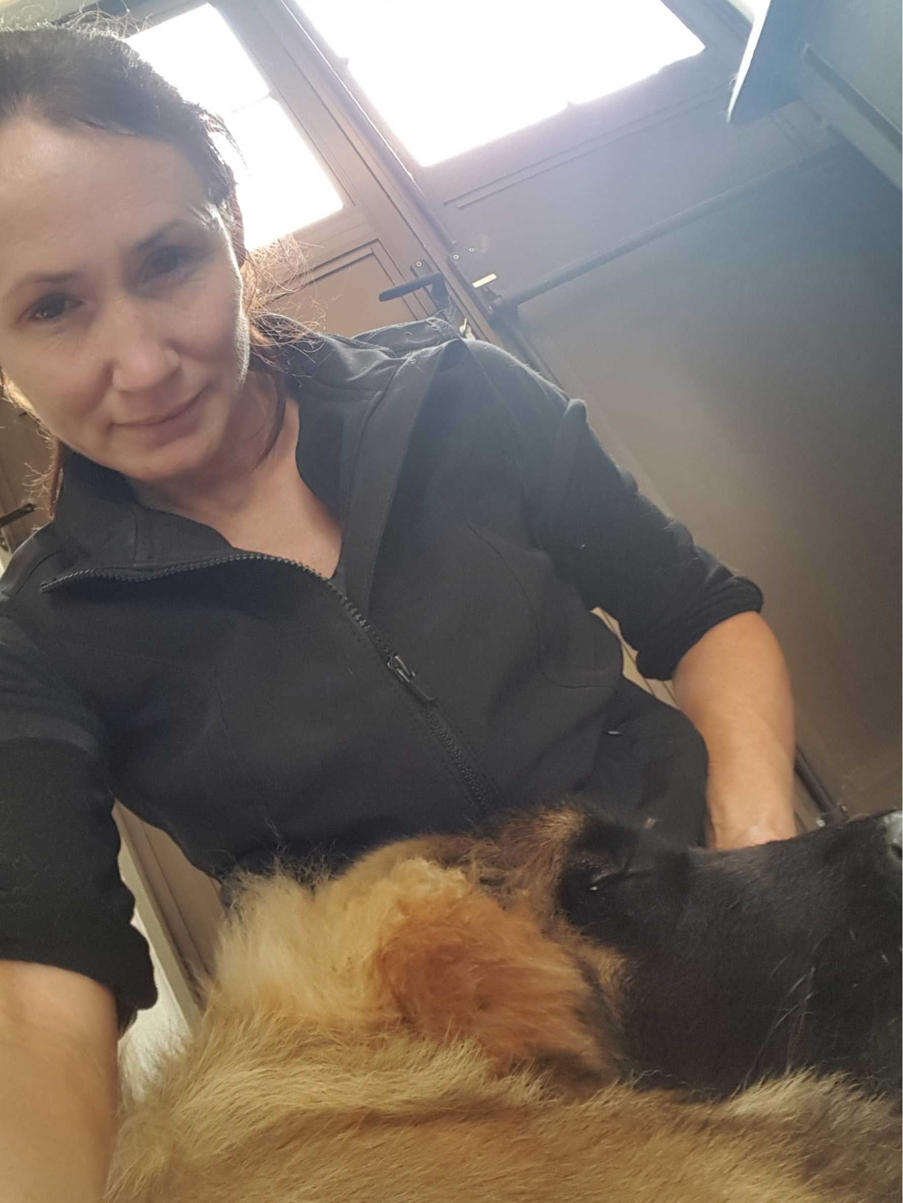 Woman in black top pets a large, long-haired dog, likely indoors. They both face the camera.