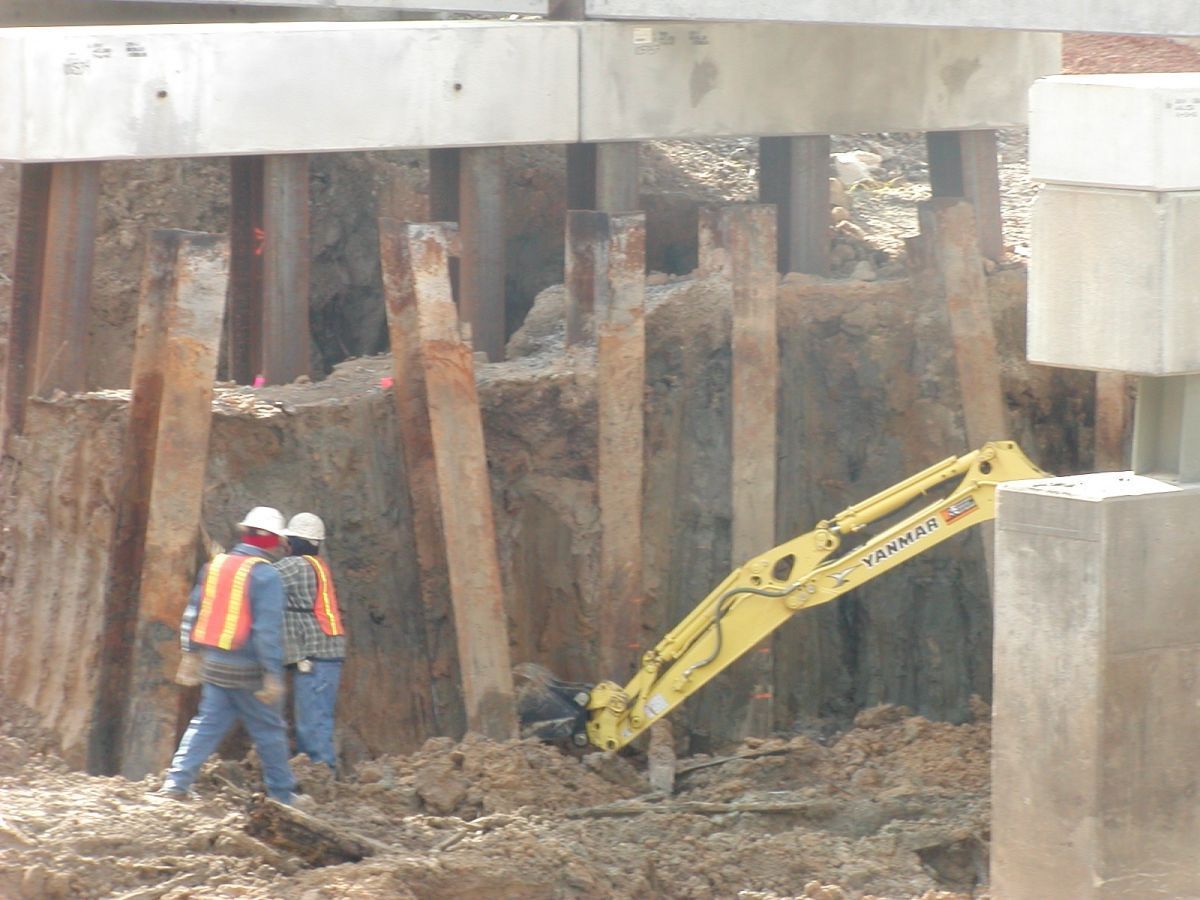 Two construction workers are standing in the dirt near a yellow excavator