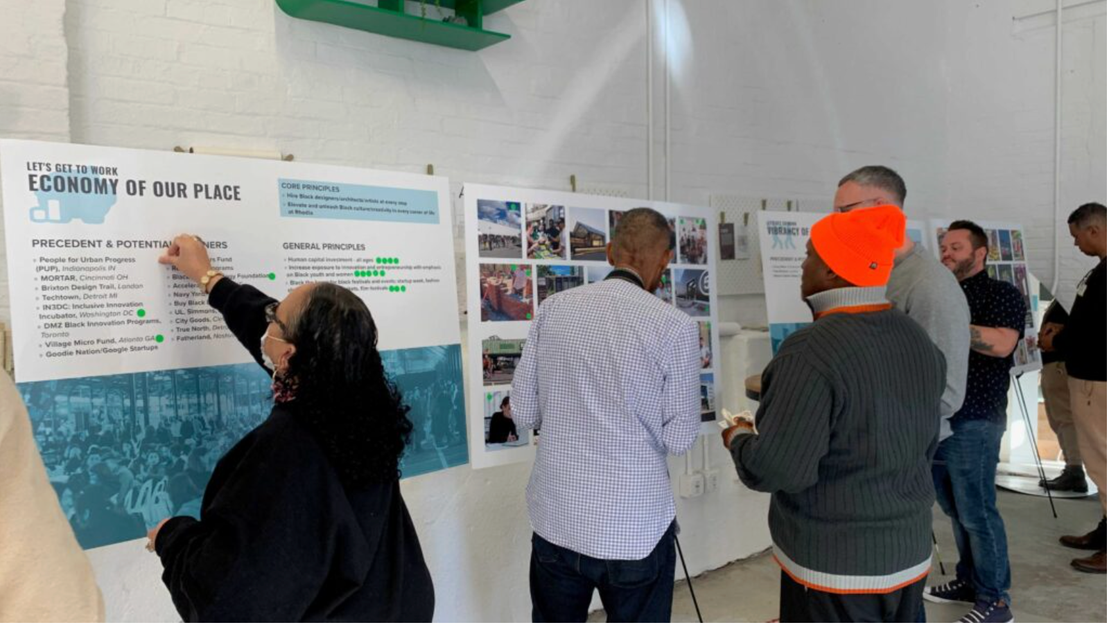 People viewing informational posters. A woman points at text, others observe. White wall, natural light.