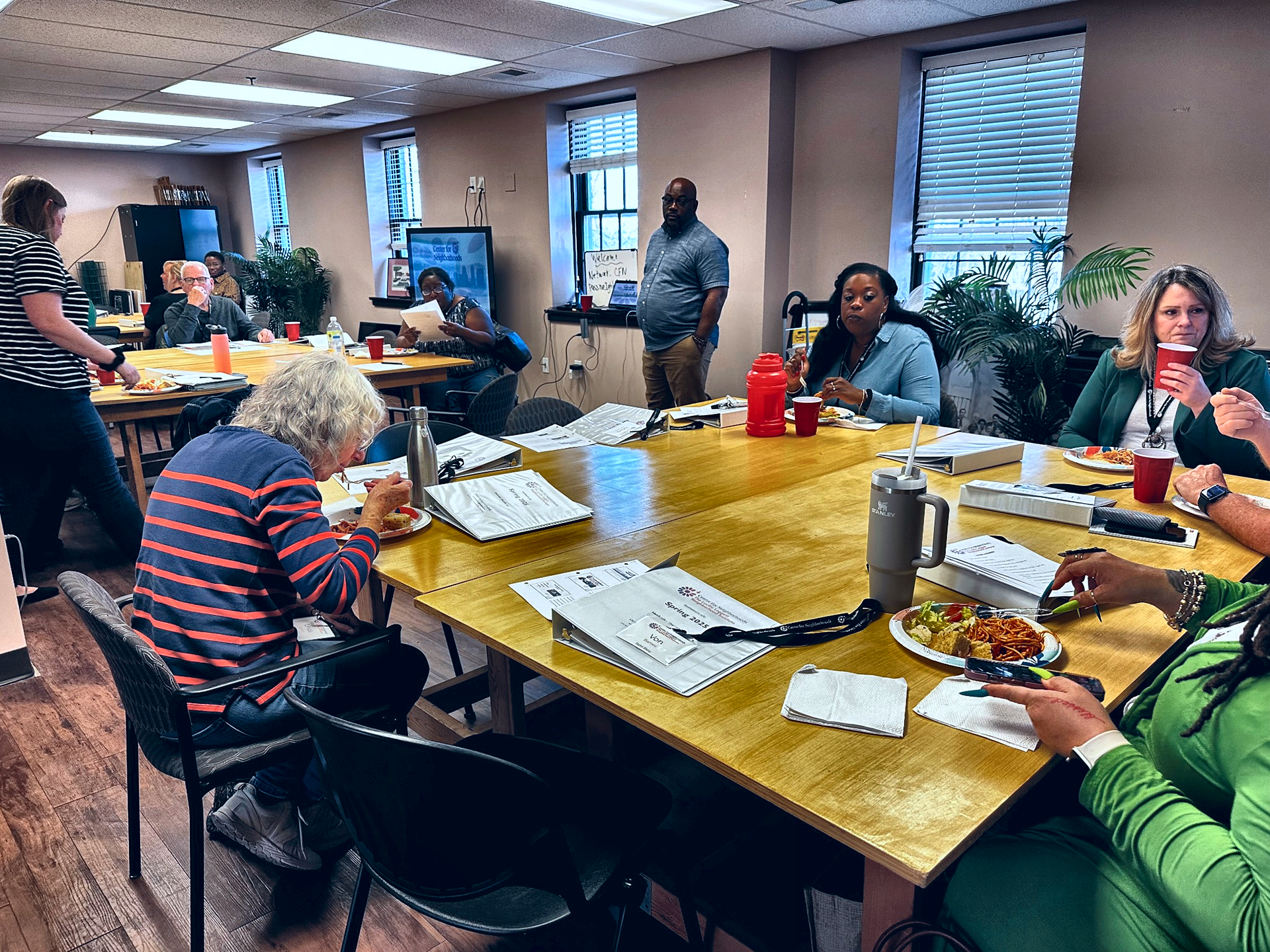 People at a table in a room, eating and working. Several papers and meals visible.