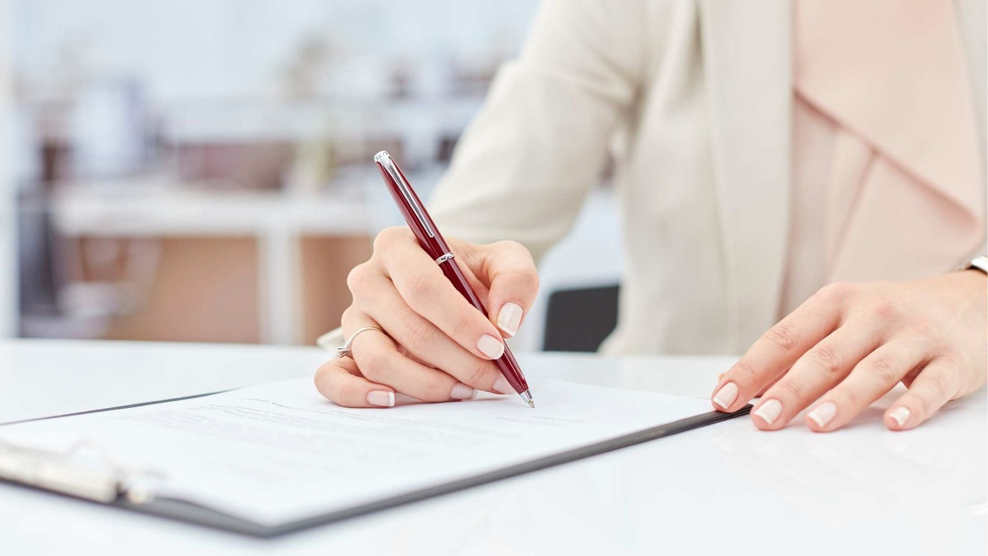 Person writing in a notebook with a pencil at a white desk in a bright office.