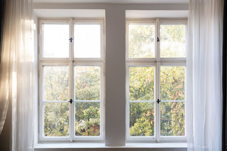 Two white-framed windows with sheer curtains, sunlight streaming in and trees visible outside.