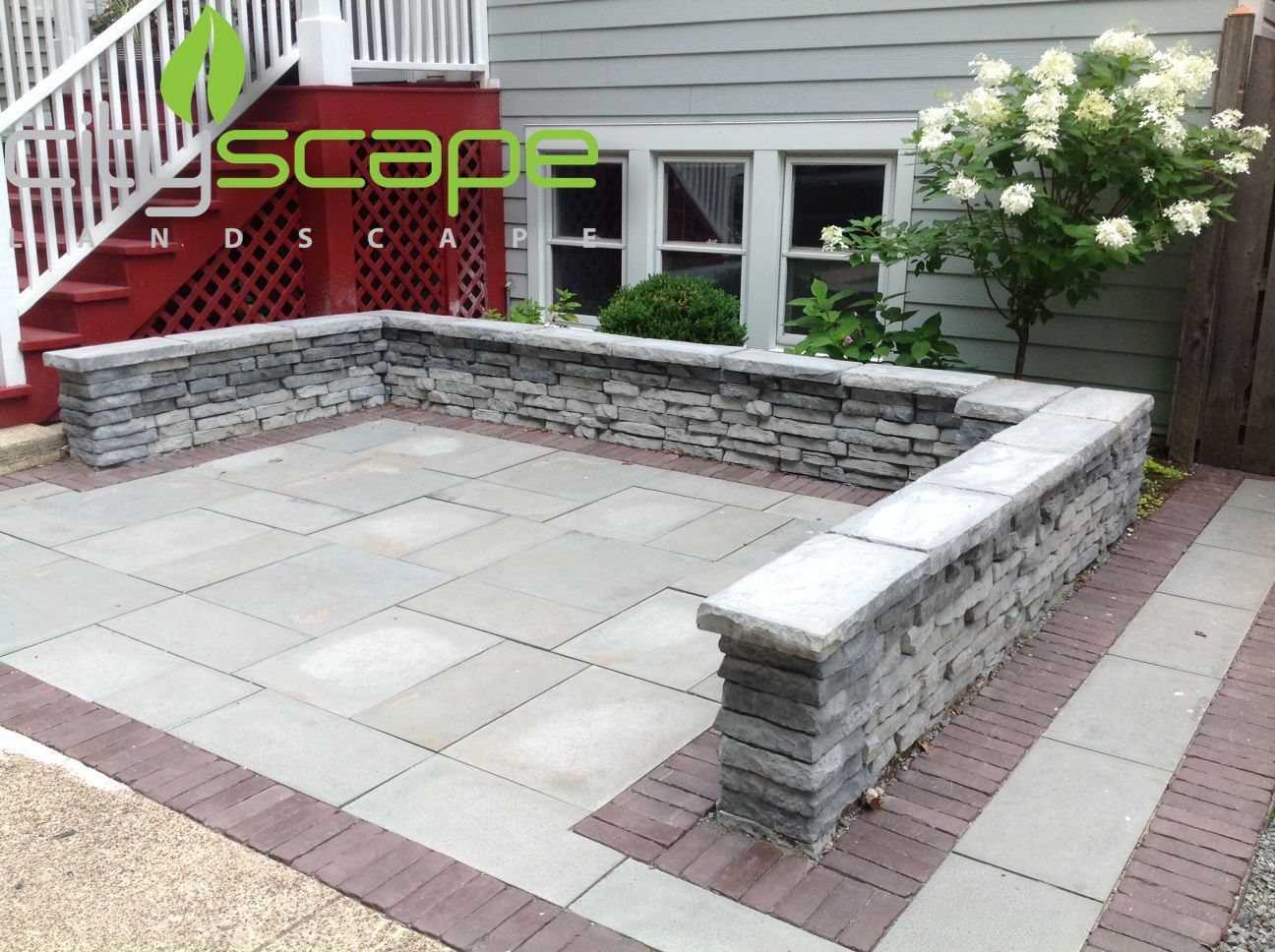 Stone patio with low retaining wall and red brick border.  Backyard setting.