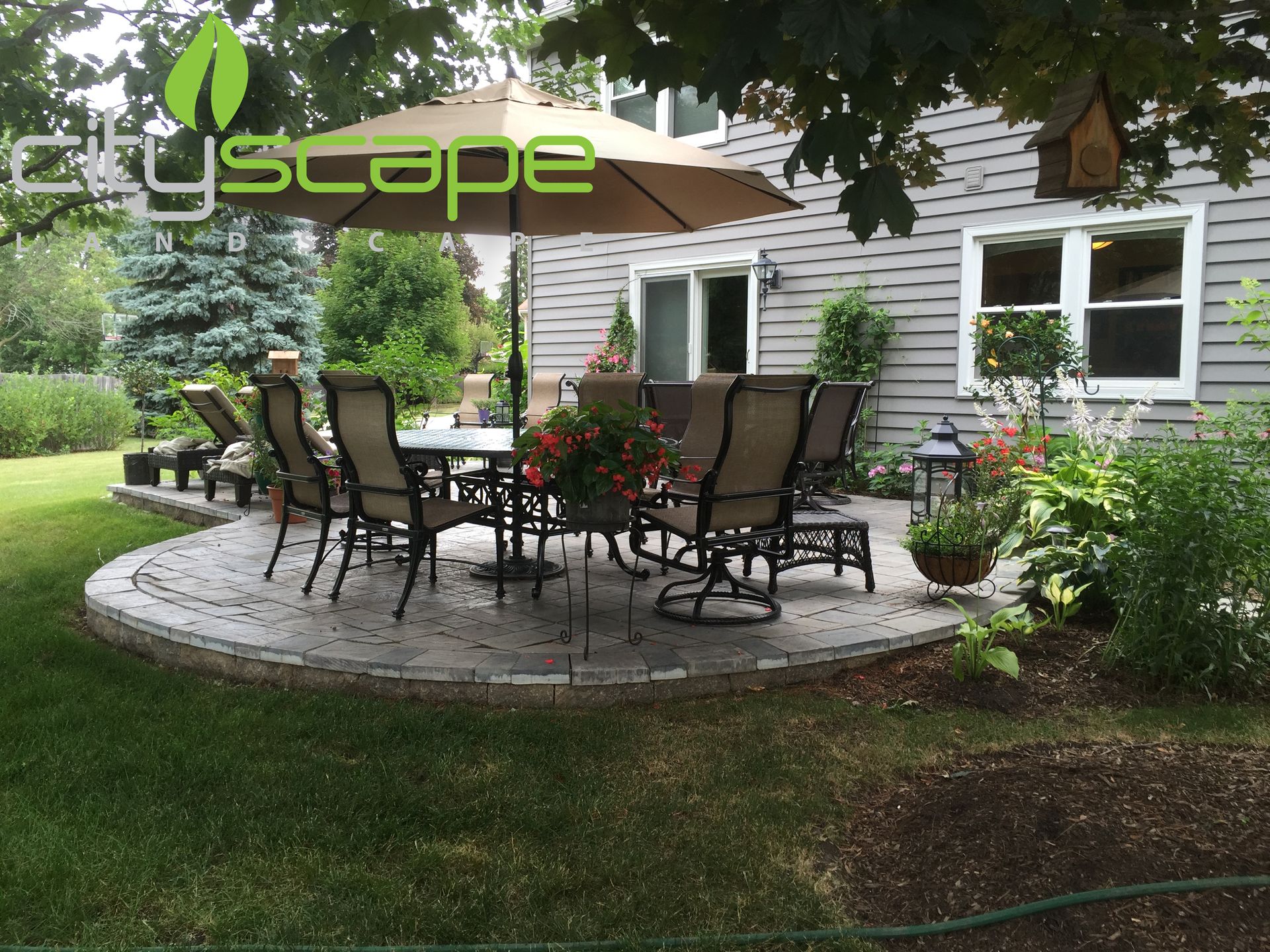 Patio with outdoor furniture, umbrella, and surrounding greenery next to a house.
