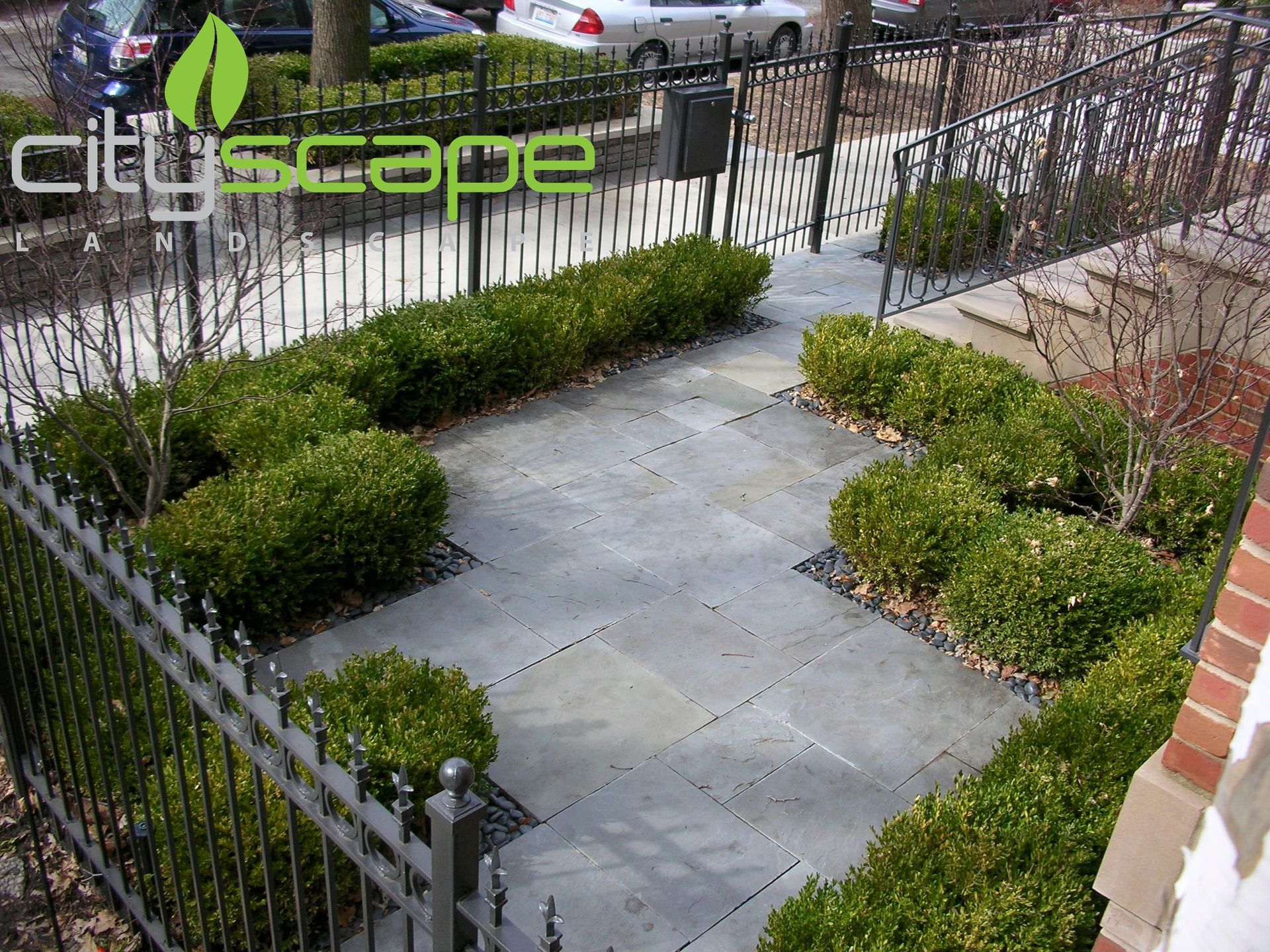 Small urban garden with hedges, paved area, and black wrought iron fence.