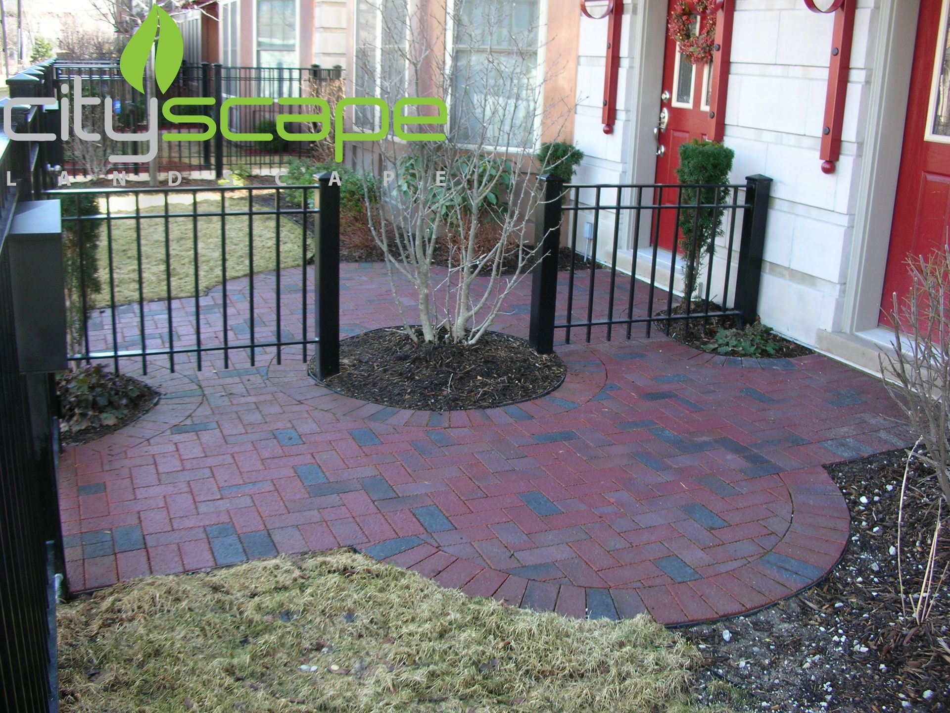 Brick patio with black metal fence and small tree.