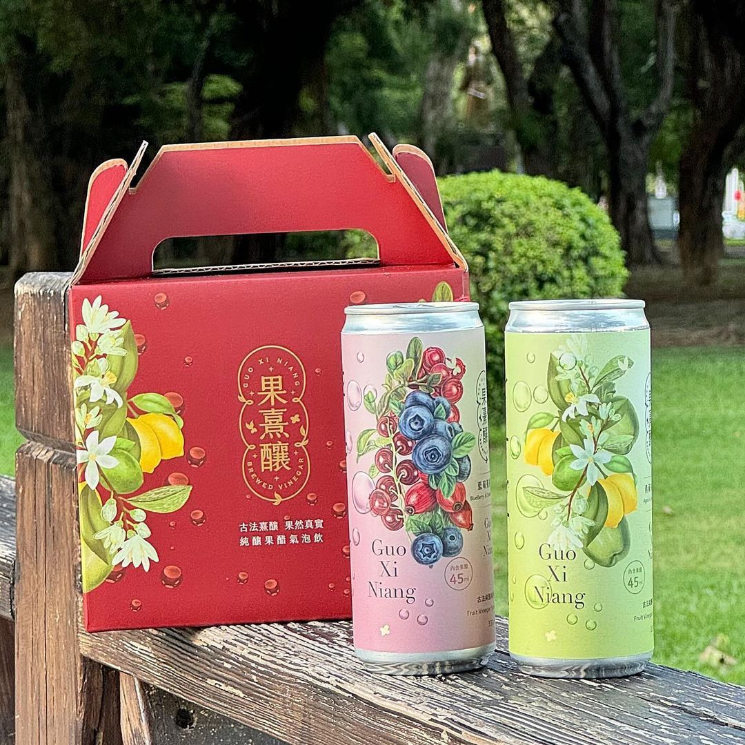 戶外木質表面上放著一個紅色禮盒和兩個飲料罐。罐子上裝飾著水彩水果和花卉圖案。