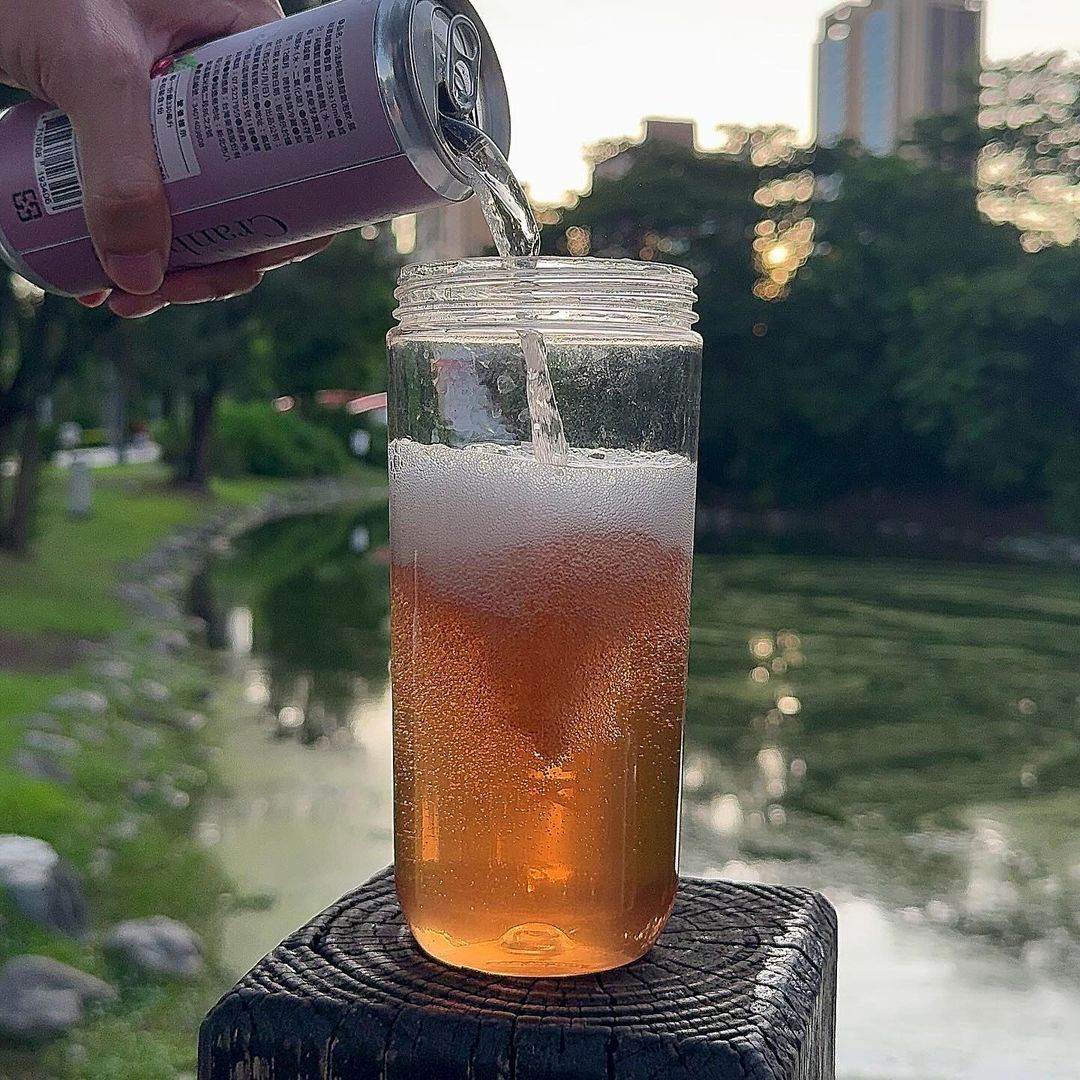 戶外，靠近水邊，一罐飲料被倒入一個高高的玻璃杯中。