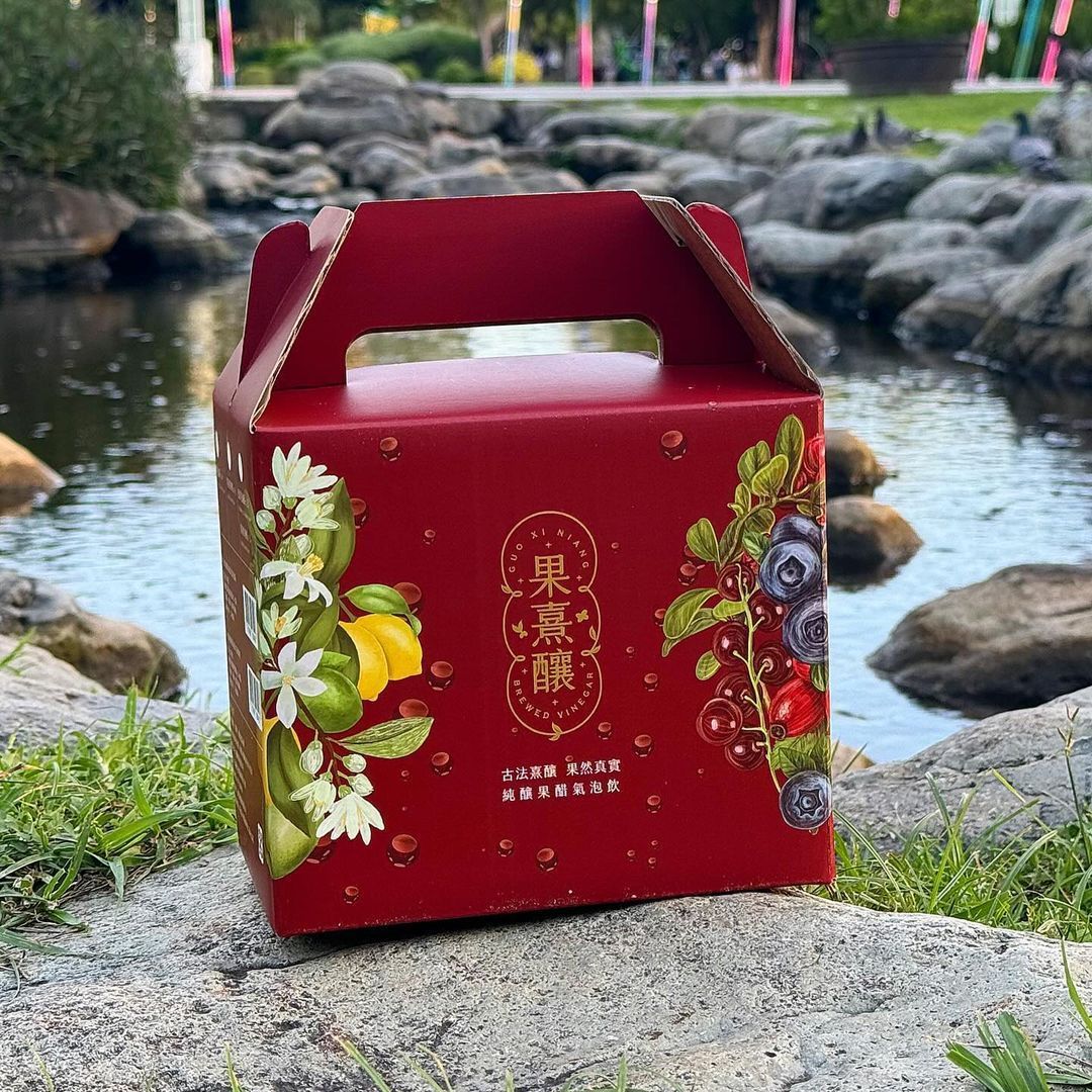 紅色禮盒，飾有水果圖案、提把和漢字，擺放在水邊。