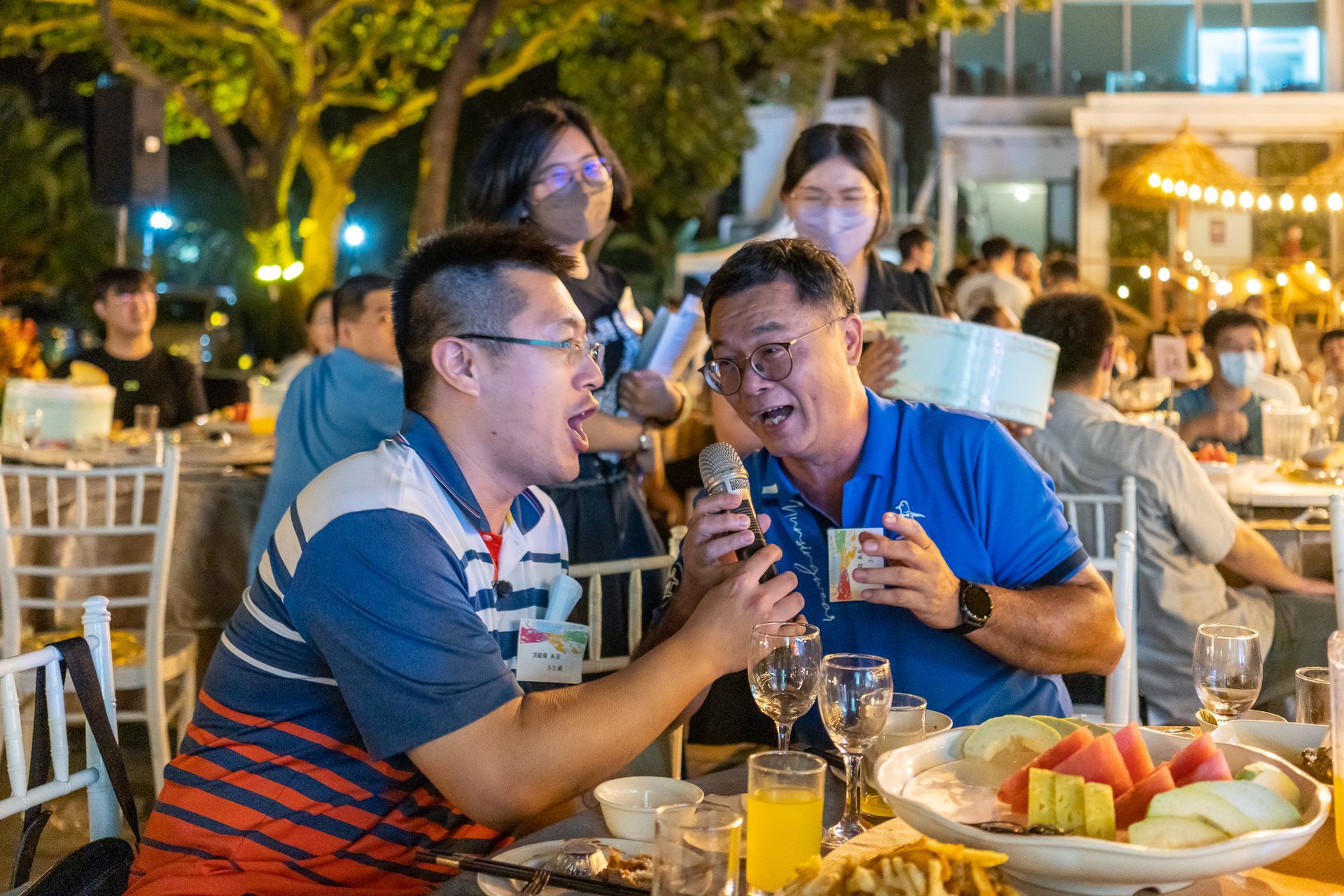 兩名男子坐在桌邊，一邊吃飯，一邊對著麥克風唱歌。