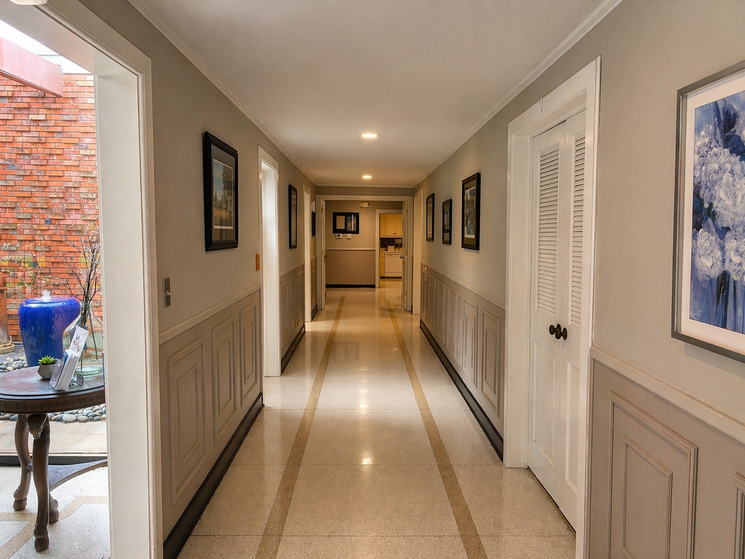 Long beige hallway with framed art, white doors, and a blue vase on a side table.
