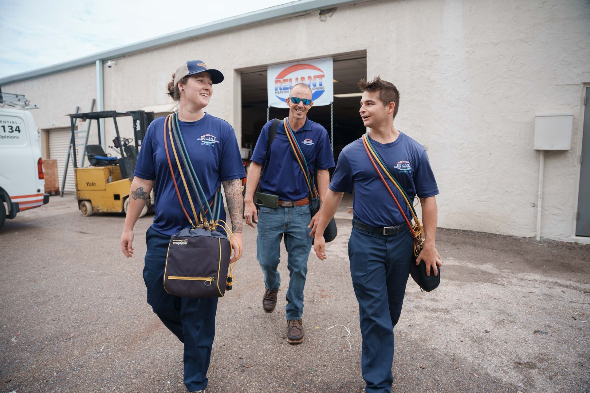 Three people in blue shirts walk past a warehouse, carrying bags and wearing caps.