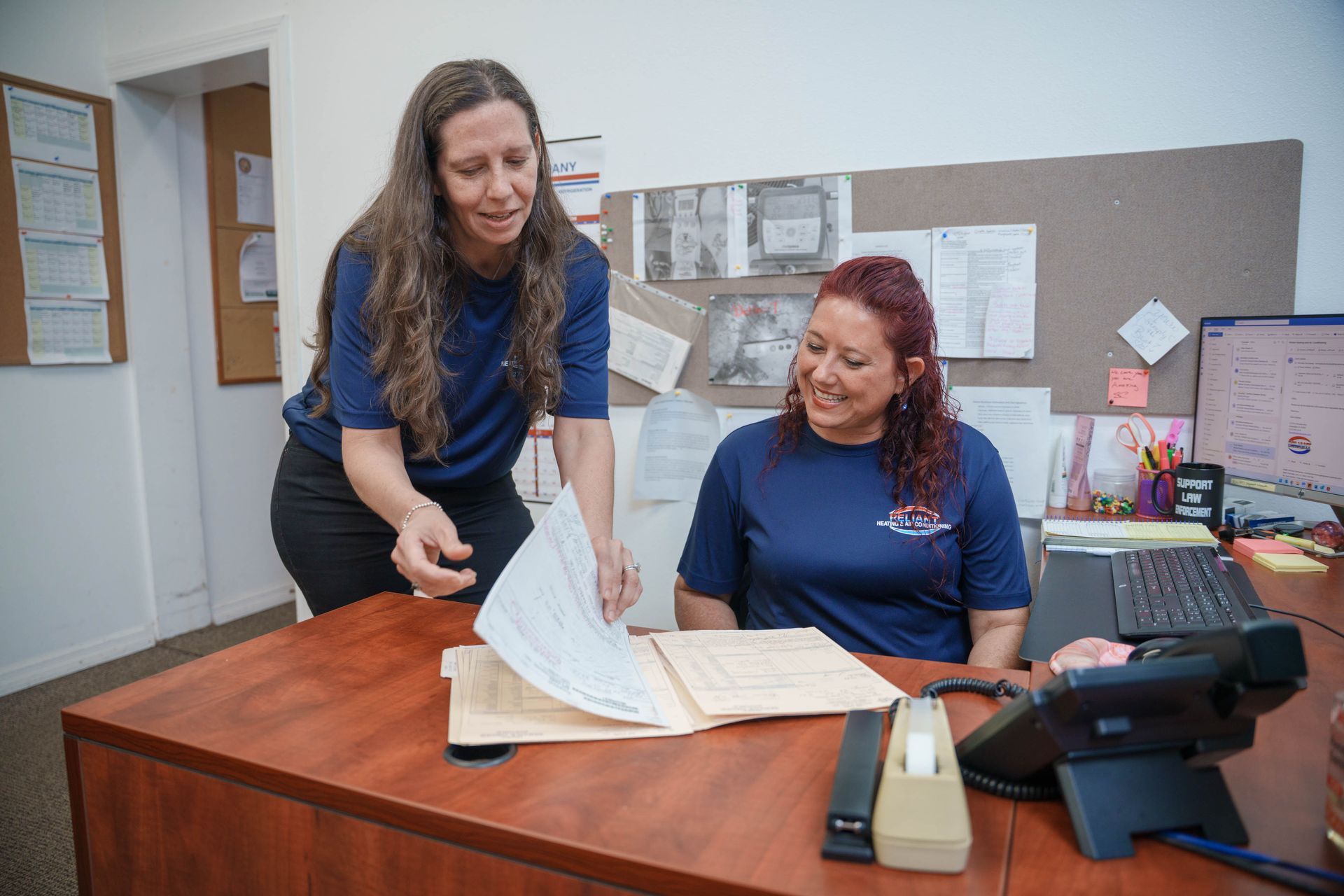 Two women review papers at a desk in an office, with files and a phone nearby.