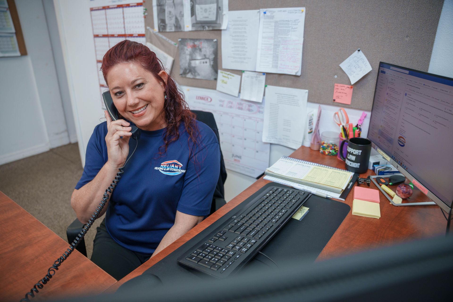 A Reliant Heating & Air Conditioning employee is sitting at a desk talking on a phone while using a computer.