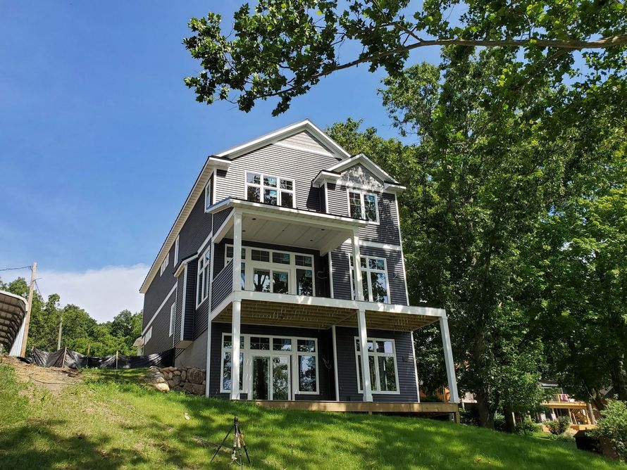 Three-story modern house with dark siding, white trim, and multiple balconies on a grassy hillside under a blue sky.