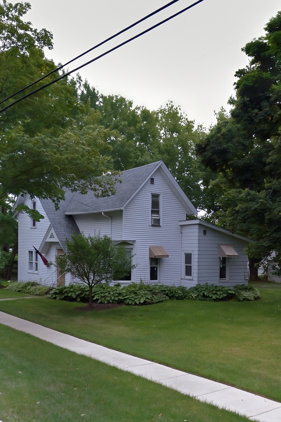 Two-story light gray house with a gabled roof and beige awnings, surrounded by greenery and a sidewalk.