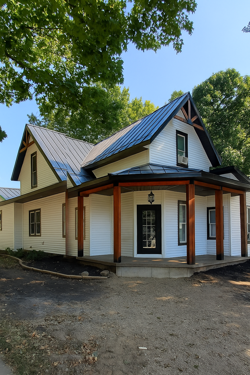 White house with black roof, wood trim, and wraparound porch. Trees in background.