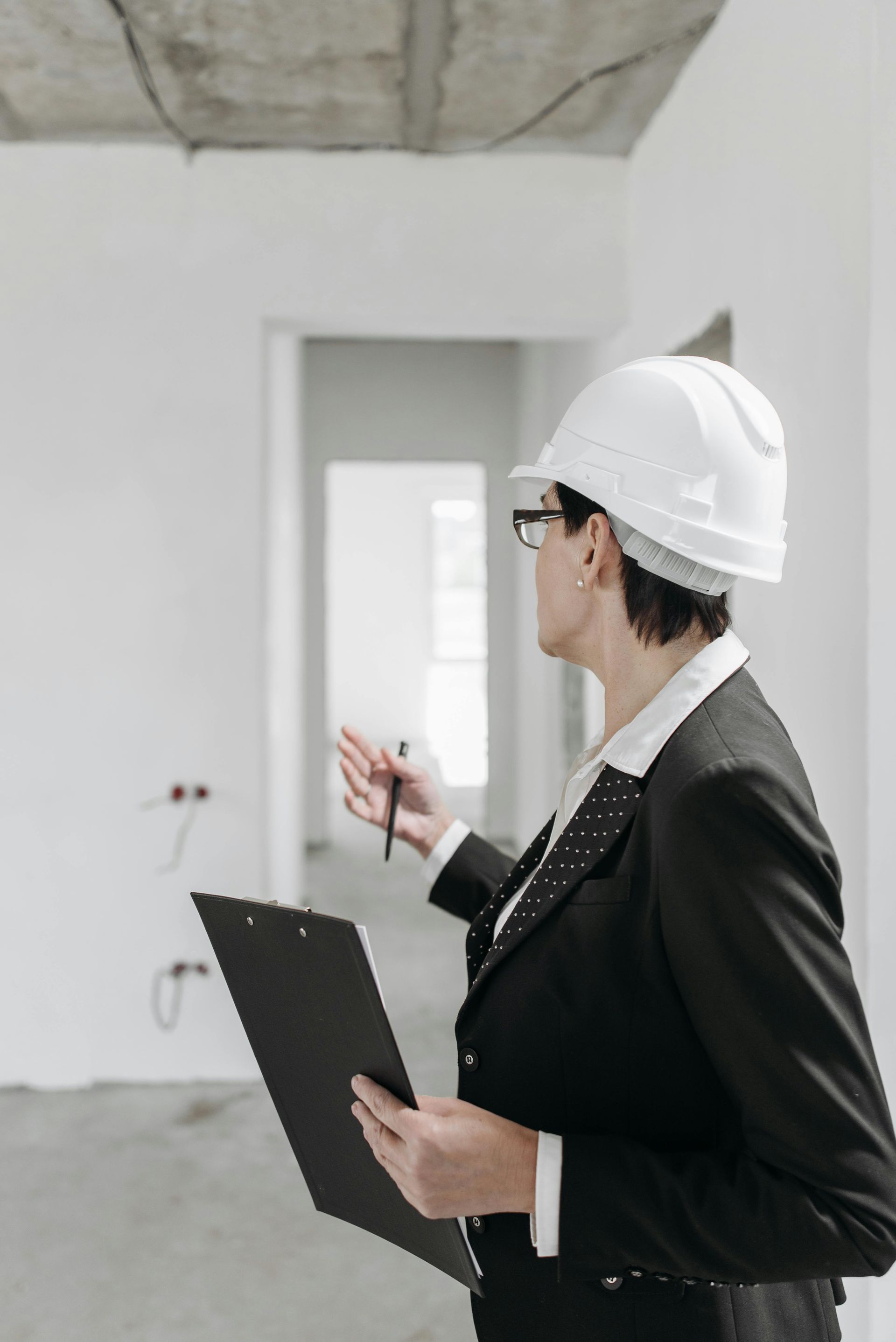 Person in a hard hat and suit inspecting a construction site, holding a clipboard and pen.