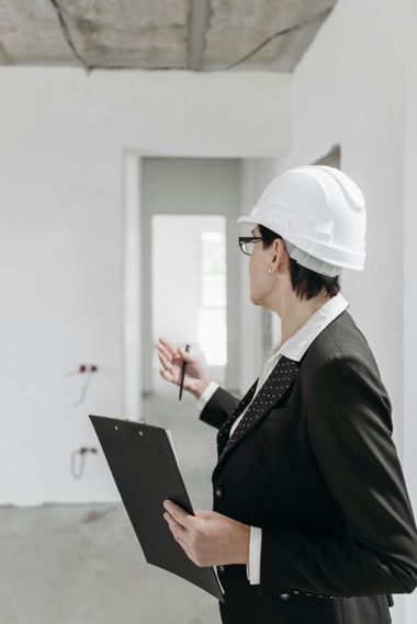 Person in a hard hat and suit inspecting a construction site, holding a clipboard and pen.