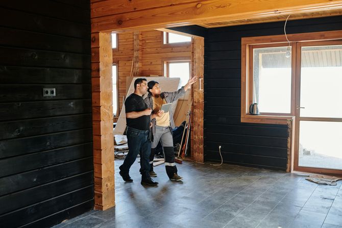 Two people in a partially renovated house; one points towards a window, the other looks on. Black walls, wood framing.