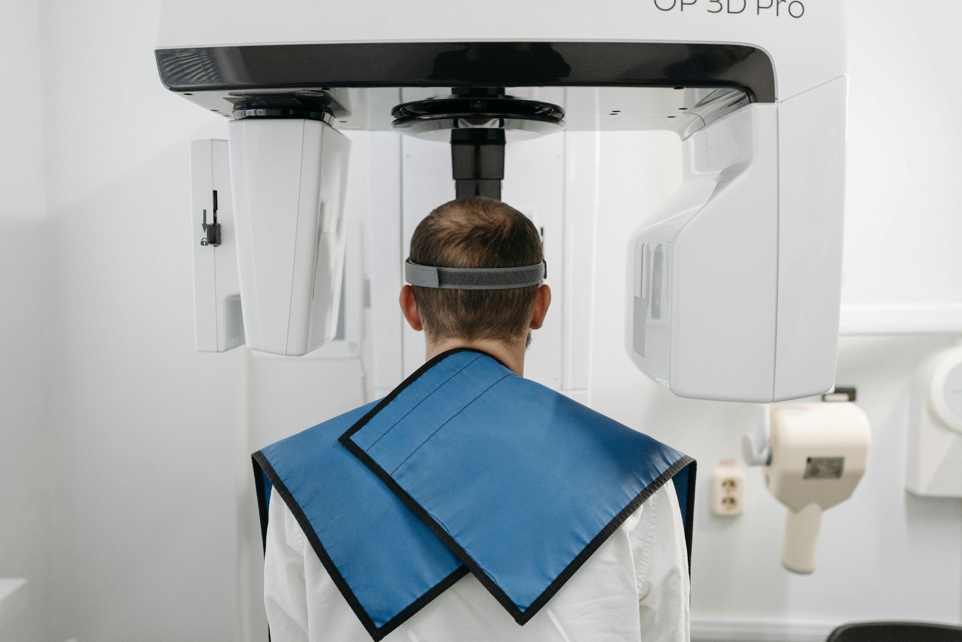 Pessoa fazendo radiografia panorâmica dentária, usando equipamento de proteção. Aparelho branco, proteção azul. Ambiente clínico.