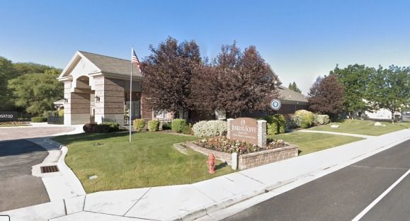 Suburban corner lot with a stone entrance sign, manicured lawn, trees, and a sidewalk along the road